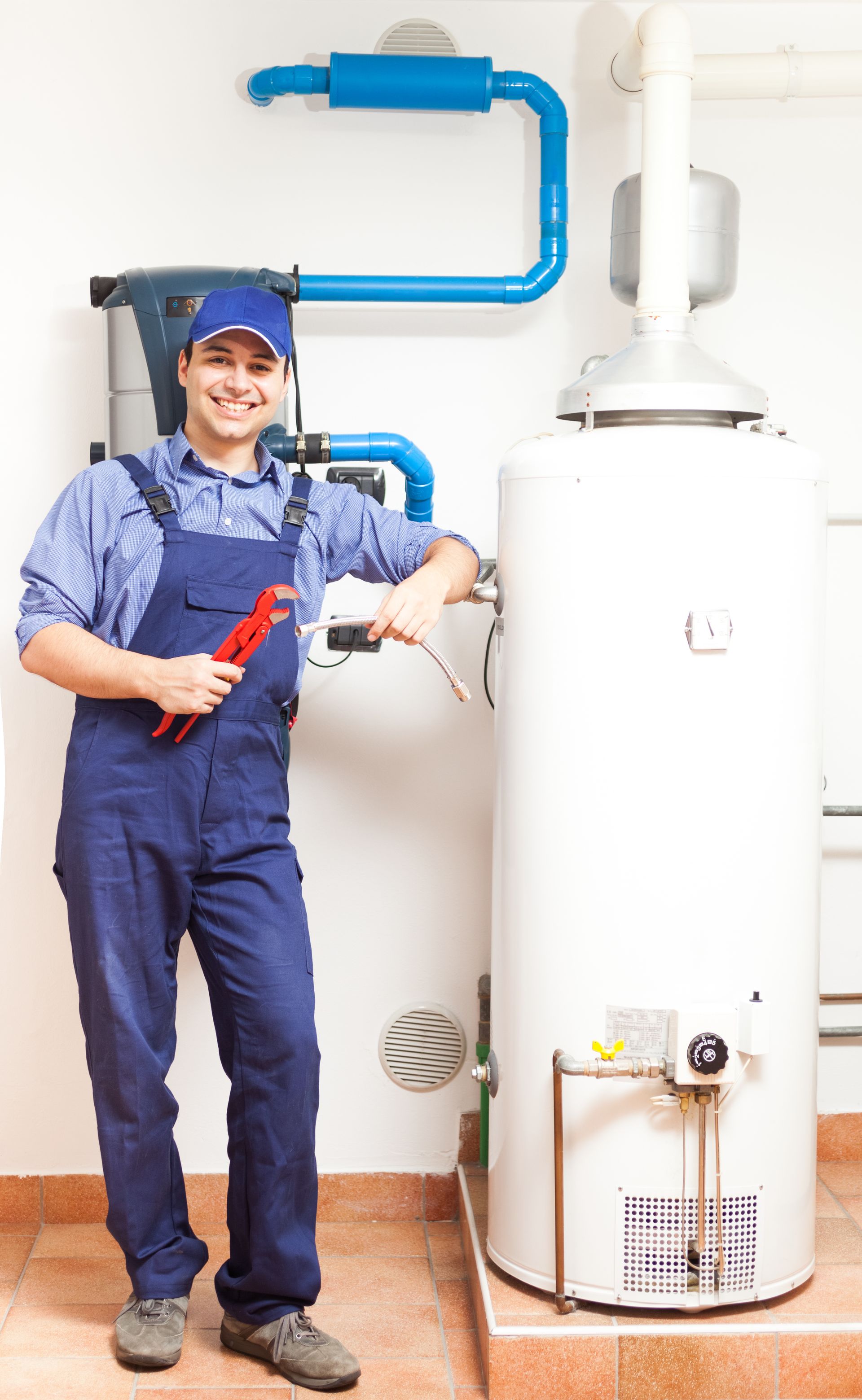Plumber in blue overalls and cap with a wrench, smiles next to a white water heater.