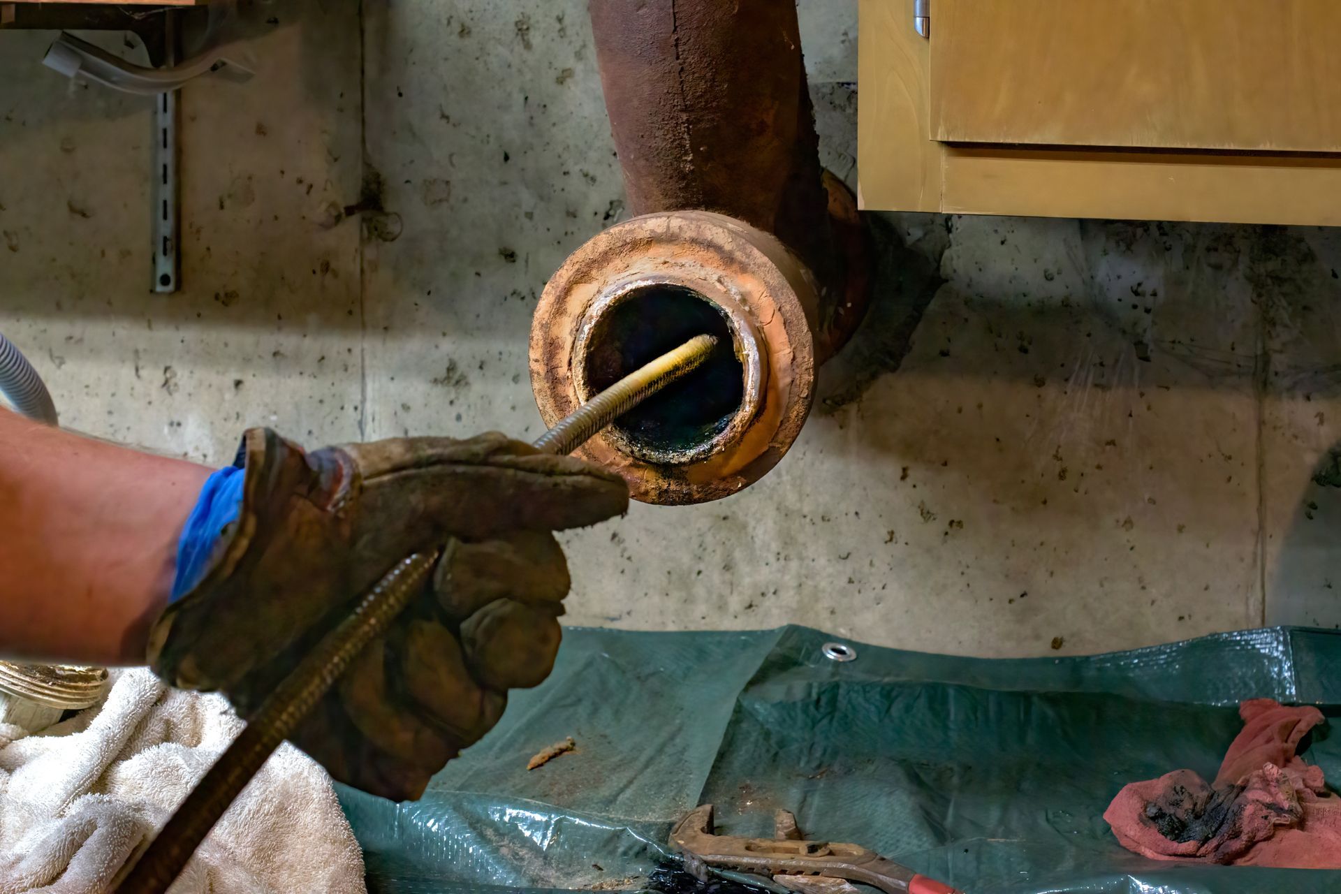 Person in work gloves cleaning a rusty pipe with a metal tool in a basement setting.