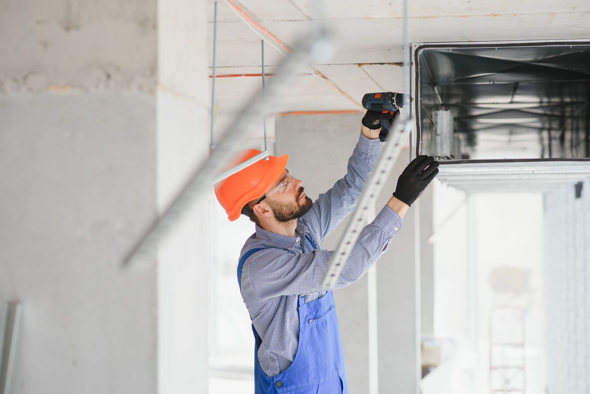 Construction worker in a blue jumpsuit and hard hat using a measuring tool on an HVAC system in a building under construction.