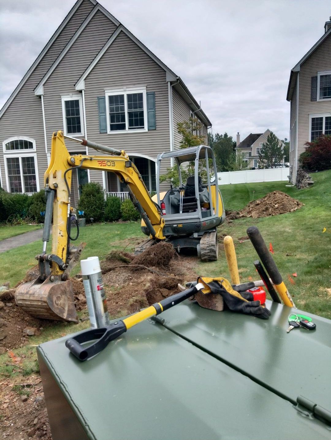 Yellow excavator digging in a residential yard near two-story houses. Shovel and tools on a green metal box.