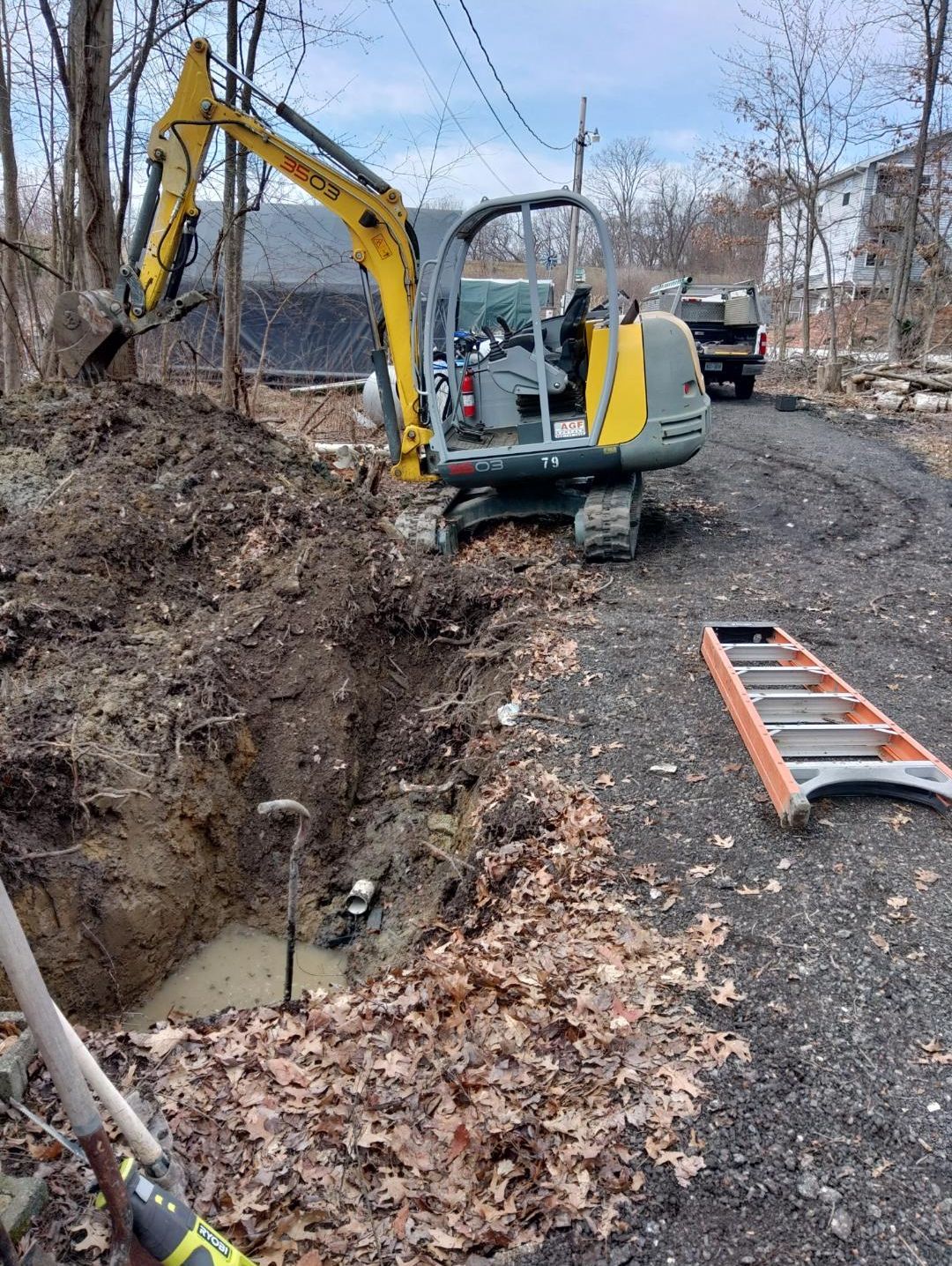 Yellow excavator digging in a muddy area next to a gravel driveway; a ladder and truck are visible.