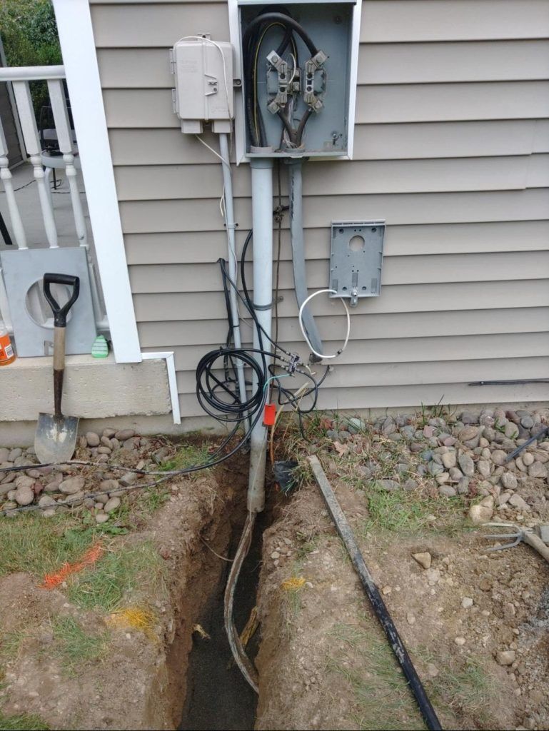 Trench dug near a house exterior with electrical wiring and boxes. Shovel and tools nearby.