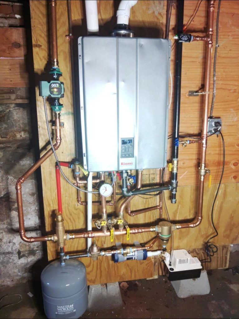 A wall-mounted water heater with copper pipes, valves, and an expansion tank, set in a wooden-walled utility room.