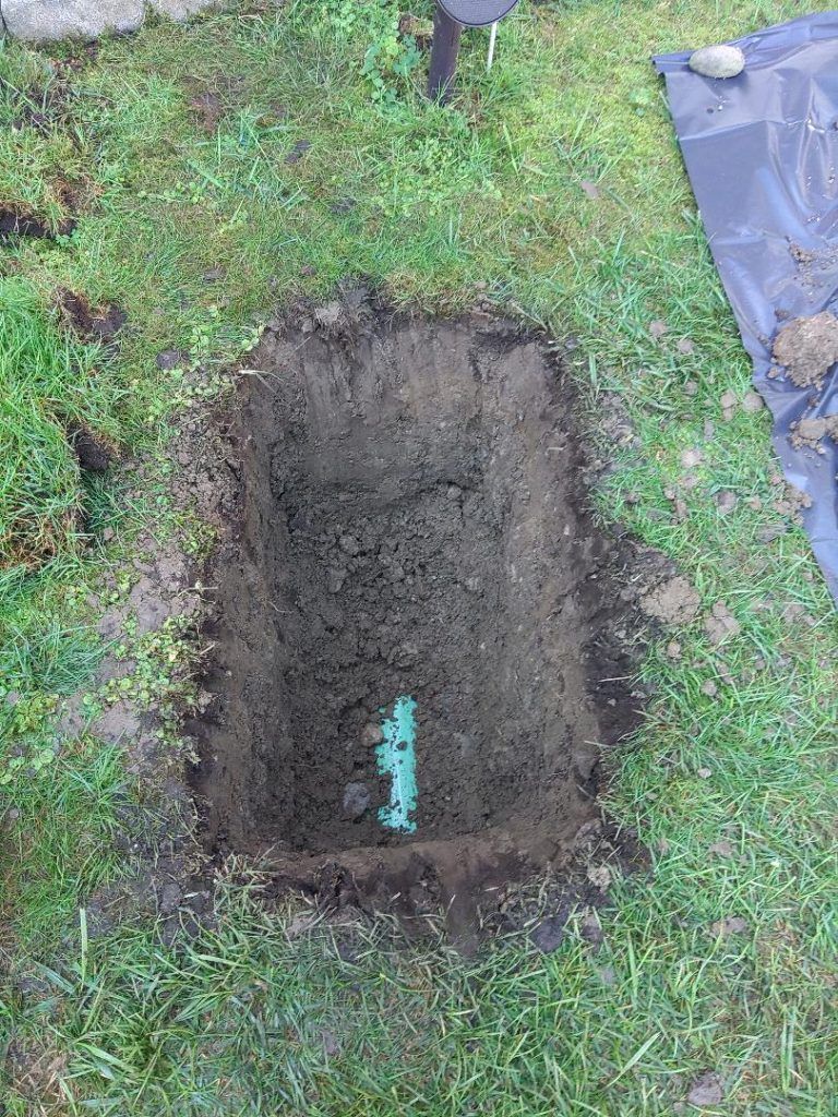A rectangular hole dug in a grassy yard, revealing green plumbing.