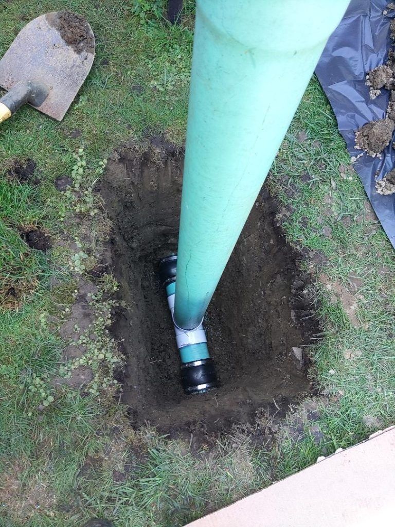 Green pipe extending from a buried junction in a square hole in grassy ground; shovel rests nearby.