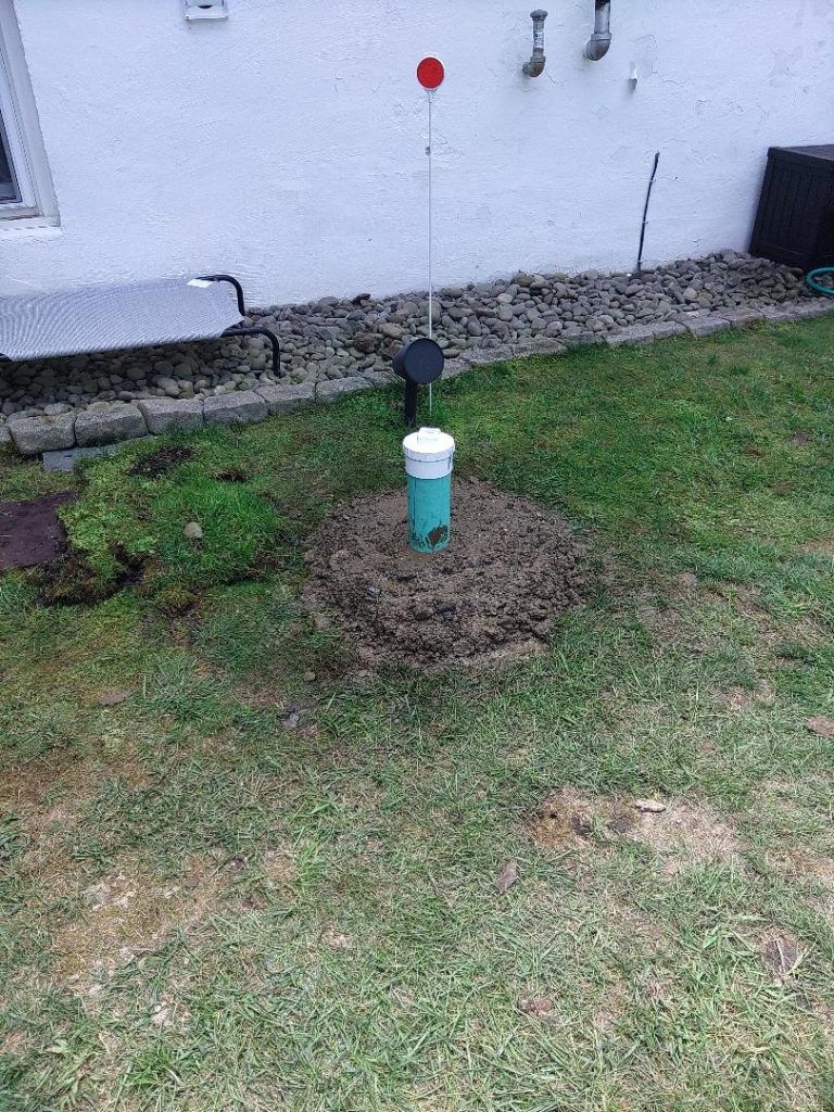 Green pipe with white cap emerging from a mound of dirt in a grassy yard, near a building.