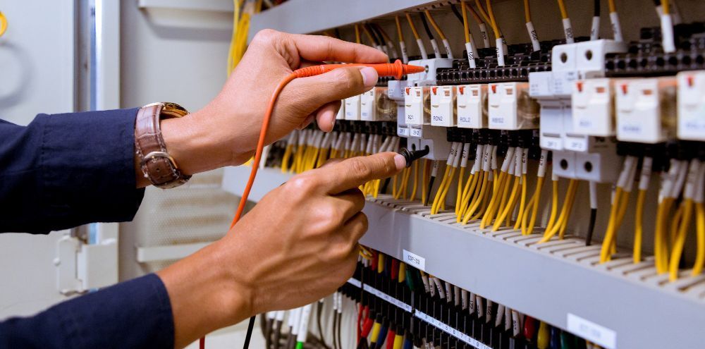 Electrician testing wires in a control panel with a multimeter. Yellow wires and white relays are visible.