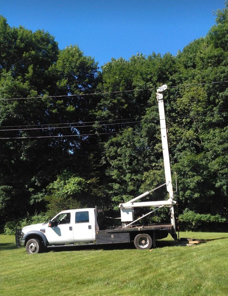 White utility truck with extended boom arm near power lines and trees on a sunny day.