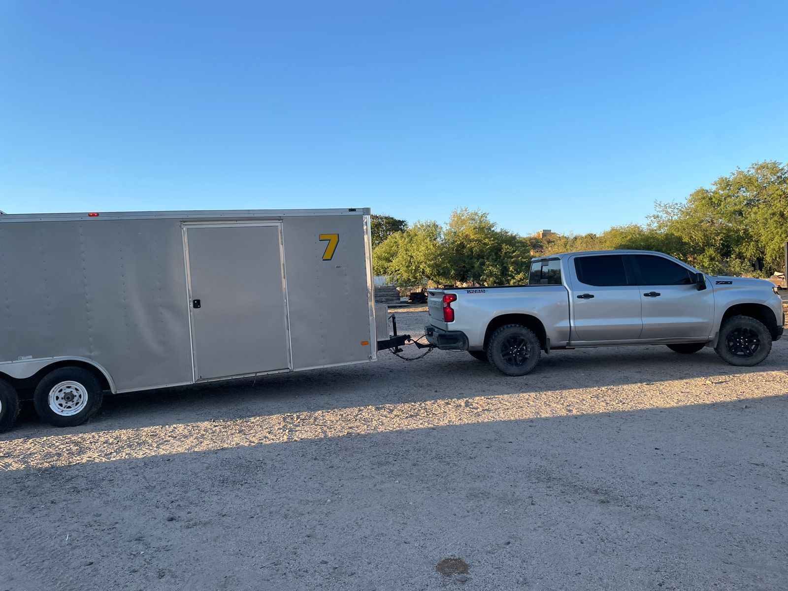 Un camión plateado arrastra un remolque en un estacionamiento de grava.