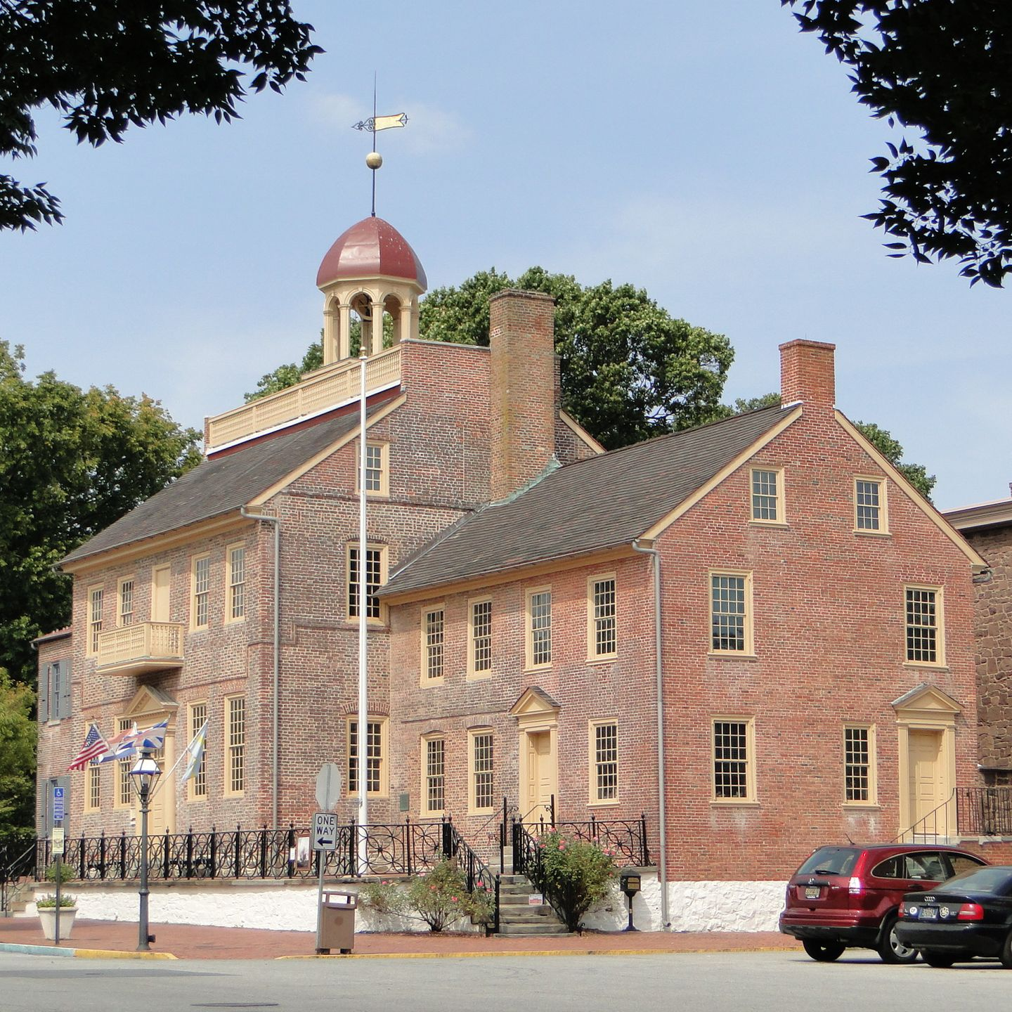 Brick building with red cupola and weathervane, in a town square, cars parked nearby.