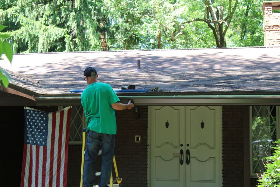 Person on a ladder cleaning a gutter on a house with an American flag.