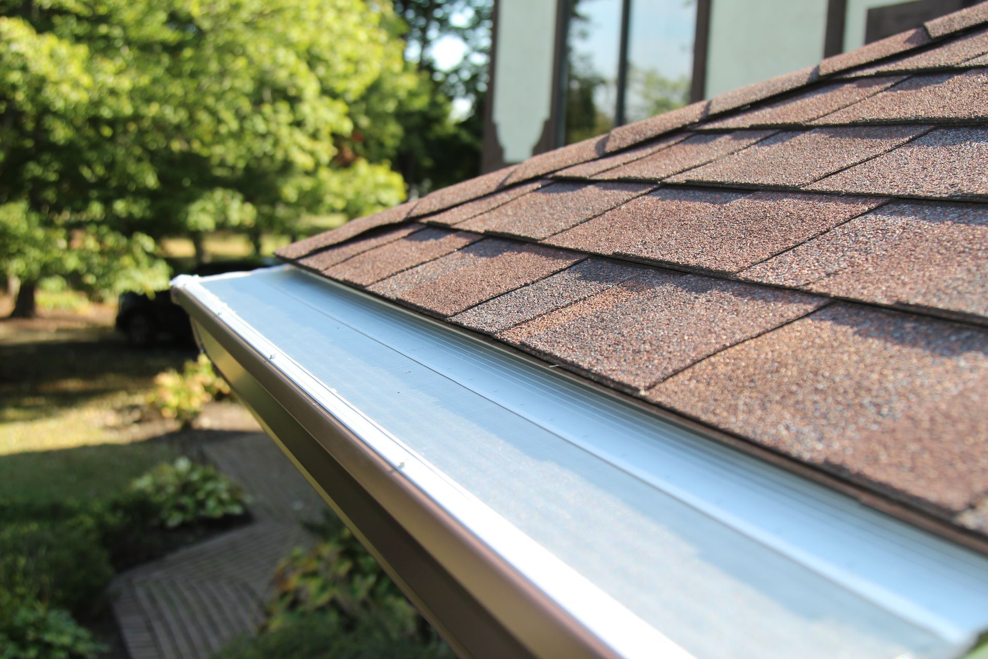 Brown roof shingles with white gutter along the edge, trees and lawn in the background.