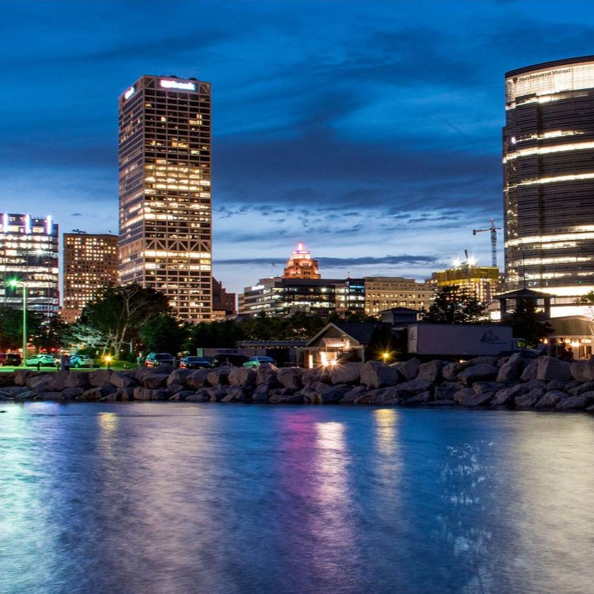 Milwaukee skyline at dusk, buildings with lit windows, water reflecting lights.