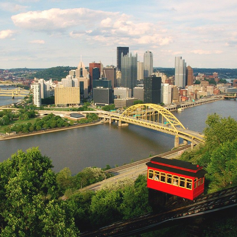 Pittsburgh skyline with a red funicular car on a hillside overlooking a yellow bridge and river.