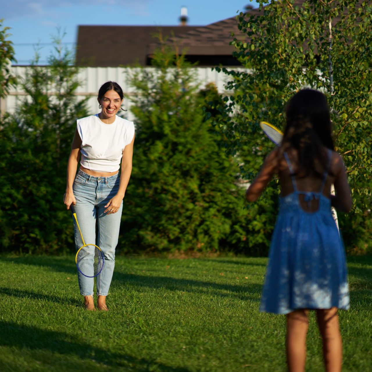 Woman smiling, playing badminton with a child in a grassy backyard. Sunny day.