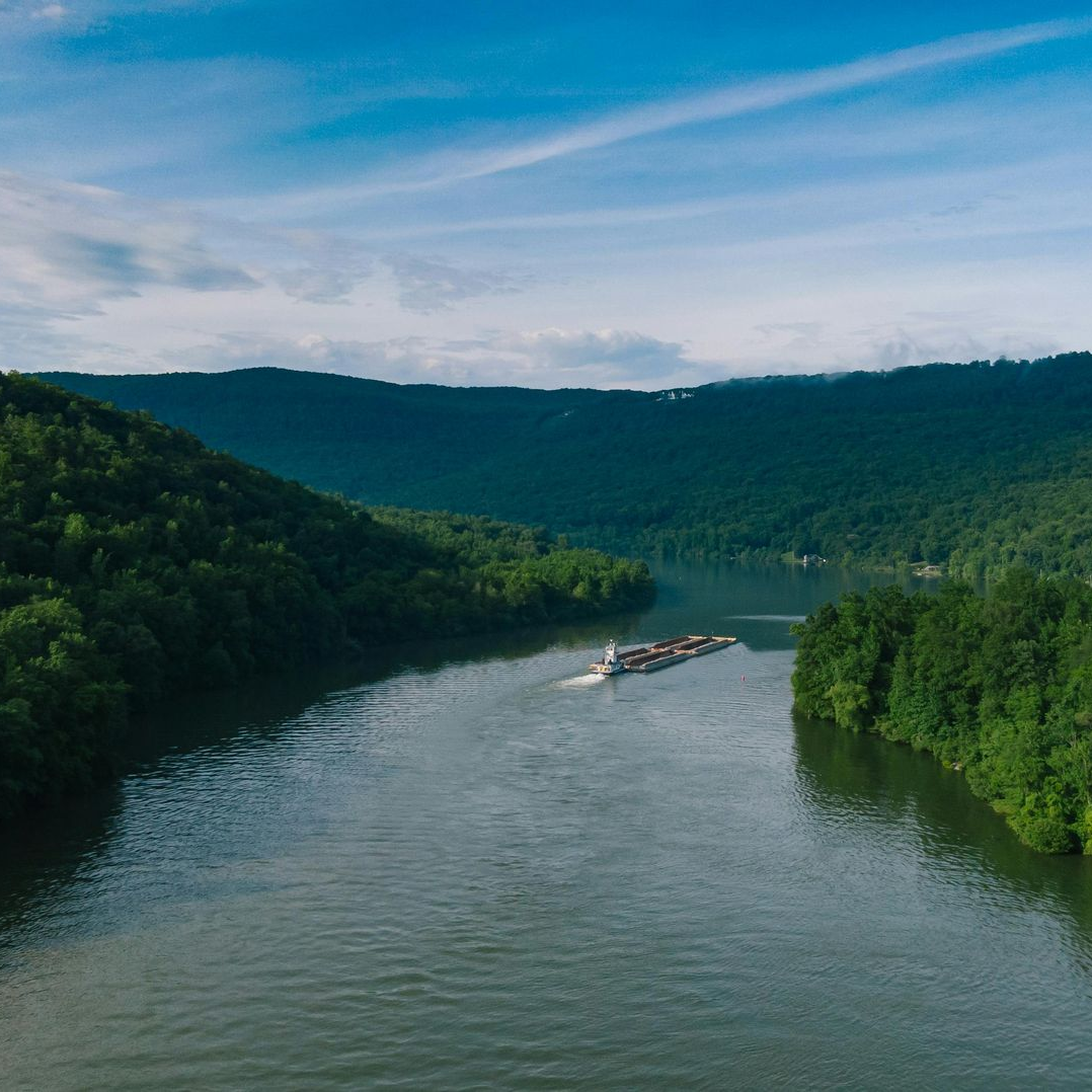 A barge travels down a wide river, surrounded by lush green forests and mountains under a blue sky.