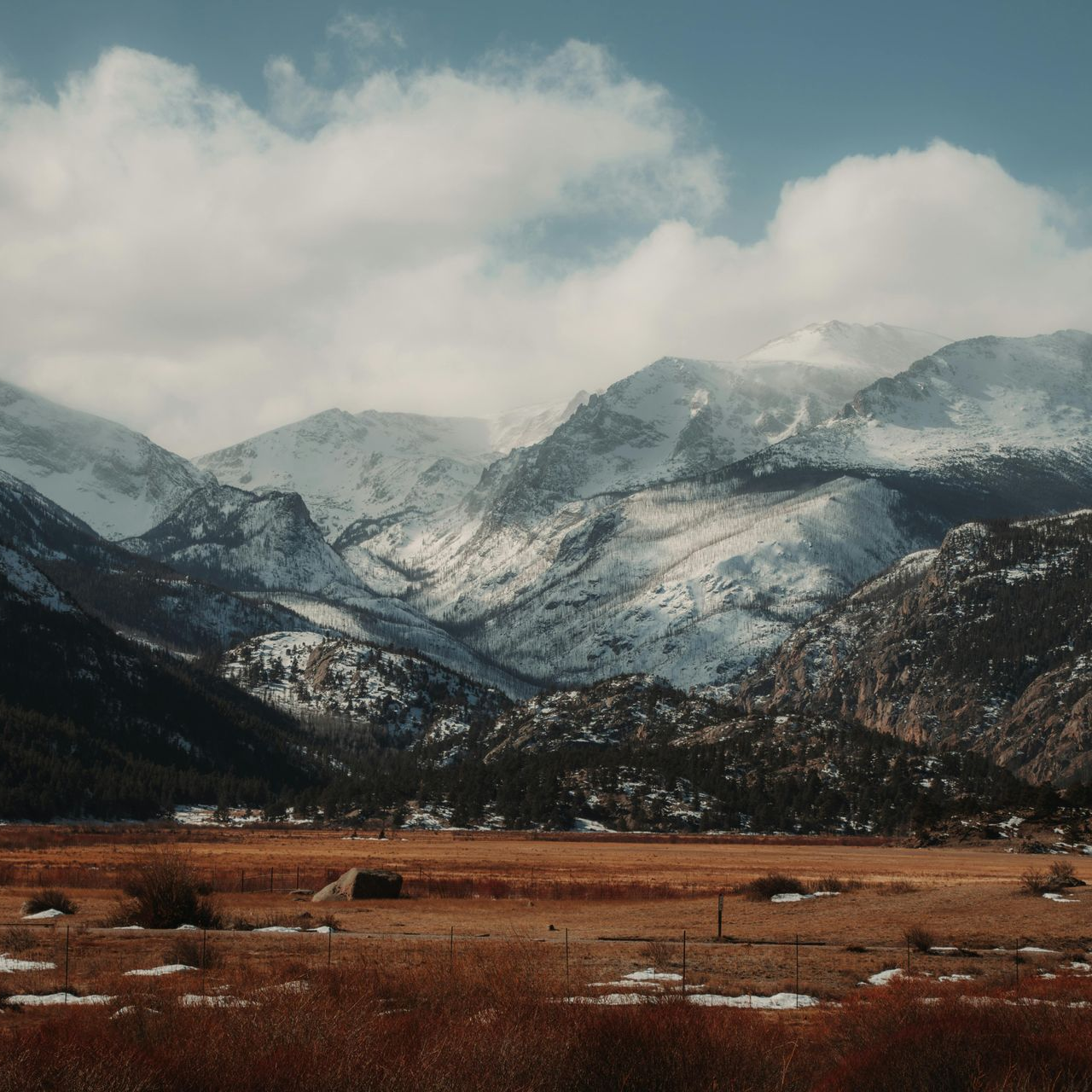Snowy mountains rise above a brown field under a cloudy sky.