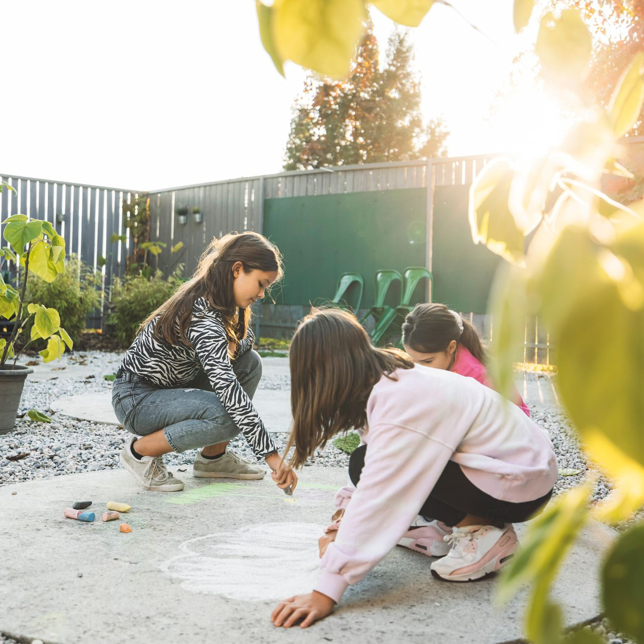 Three girls drawing with chalk on a concrete patio in a sunny backyard.