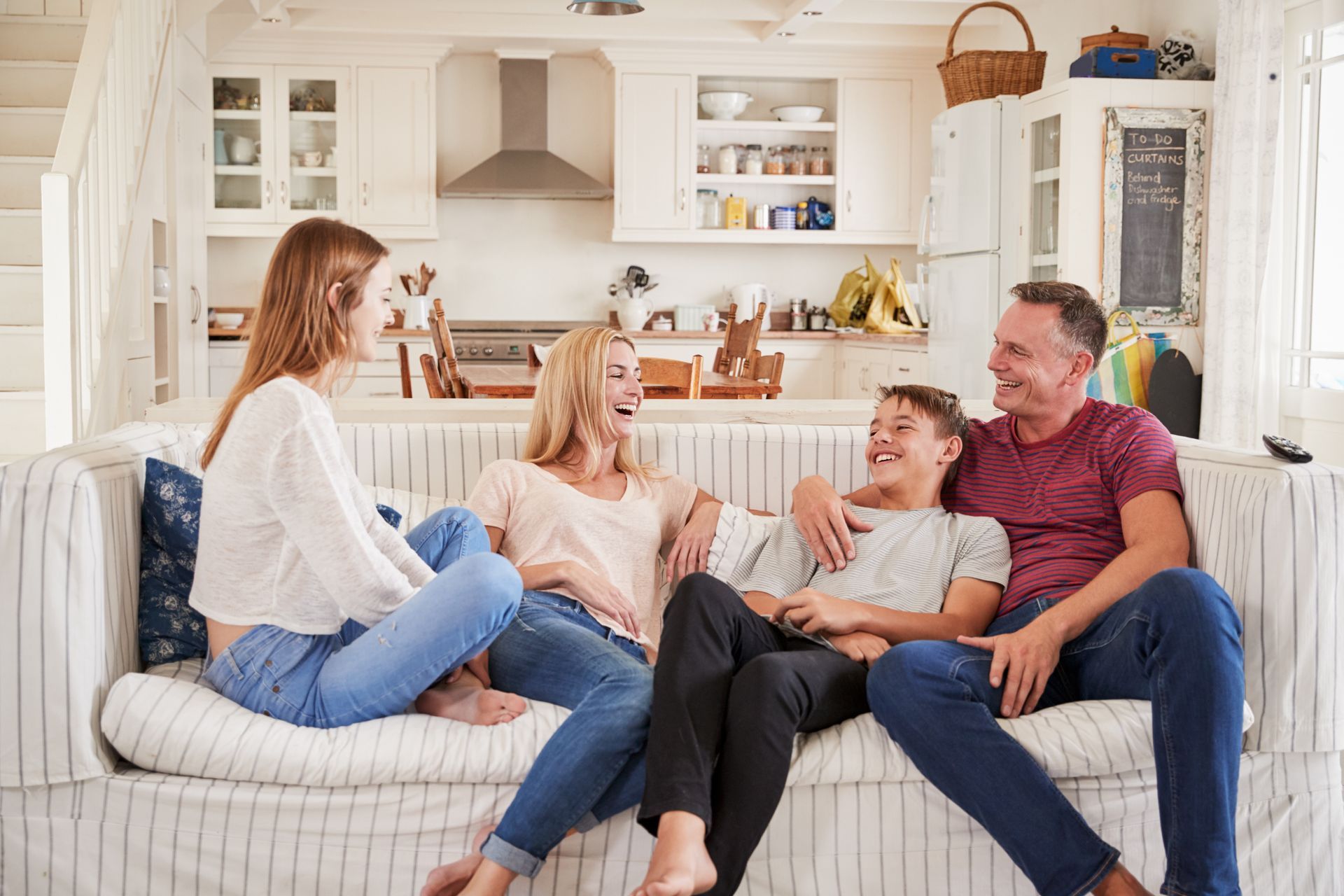 Family of four laughing together on a couch in a bright kitchen setting.