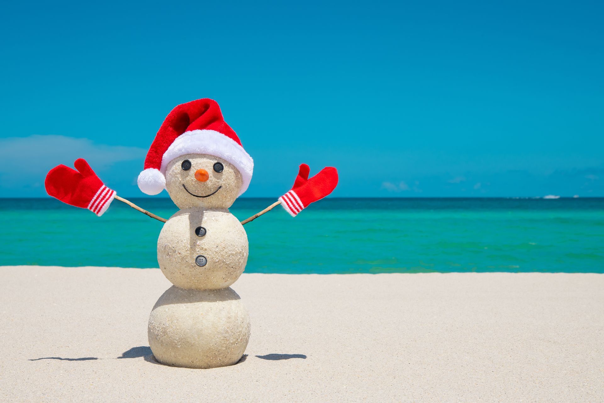 Snowman in a Santa hat and mittens on a sunny beach, arms raised, ocean and blue sky in background.