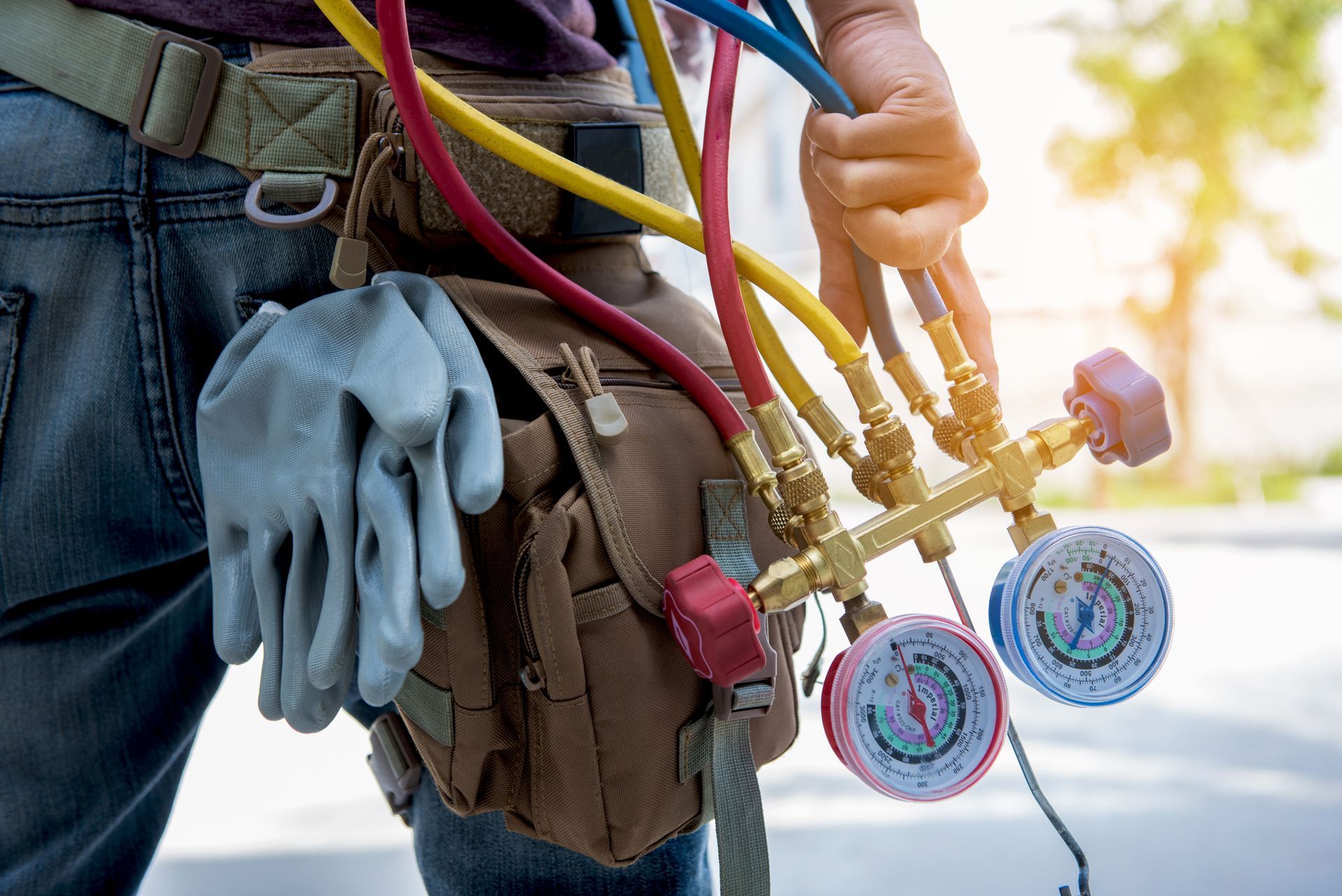 HVAC technician holding manifold gauges and hoses clipped to a tool belt.