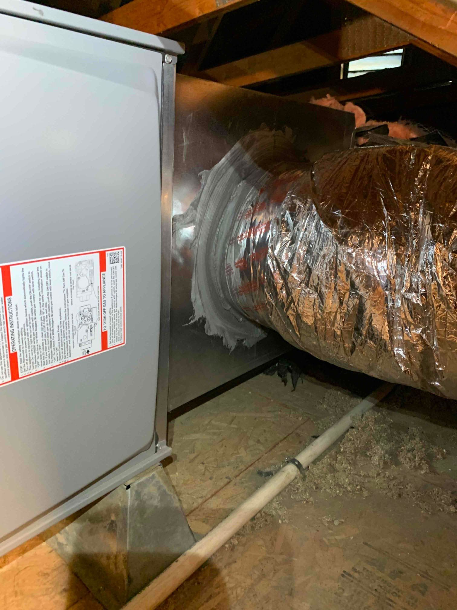 Gray HVAC unit with silver ductwork in an attic, with wooden beams and insulation.