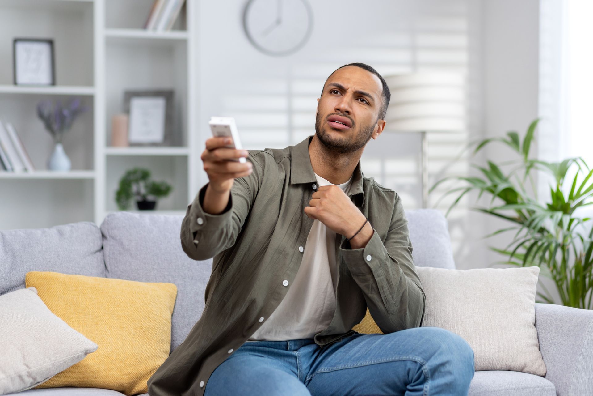 A man suffering from heat is sitting on a sofa holding a remote control to turn on an air conditione