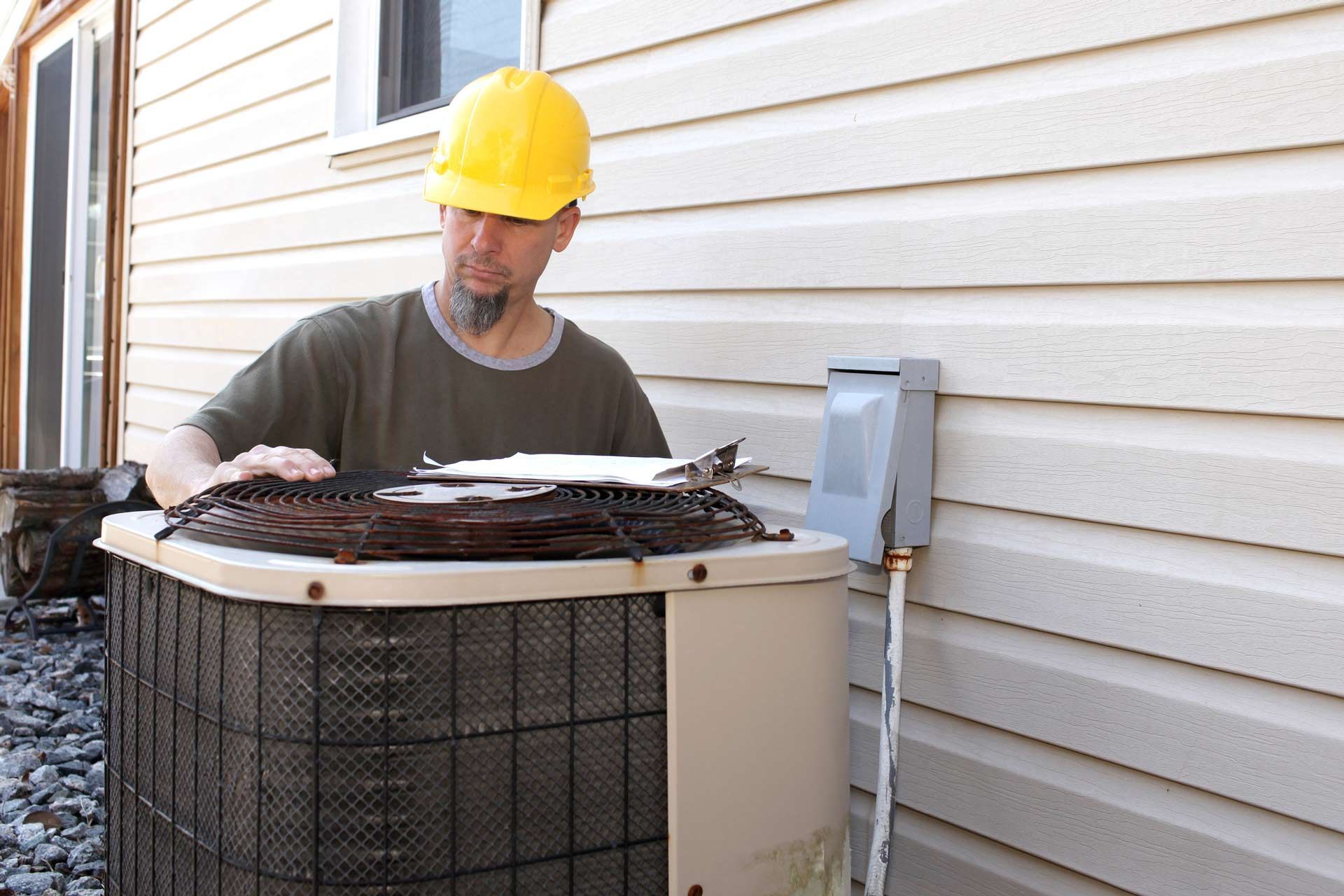 A man wearing a hard hat is working on an air conditioner outside of a house.