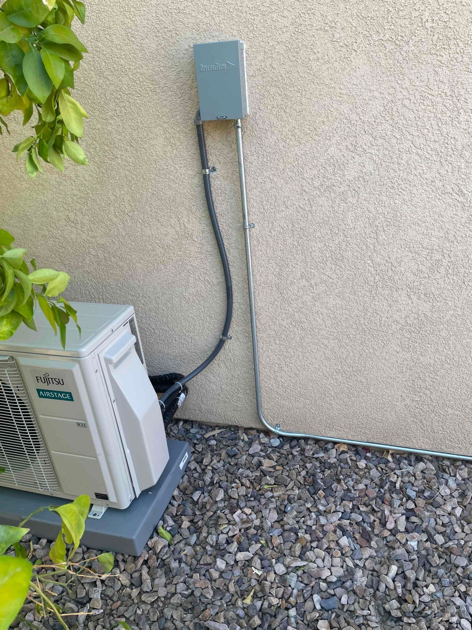 Fujitsu AC unit next to a stucco wall with electrical box and conduit on gravel.
