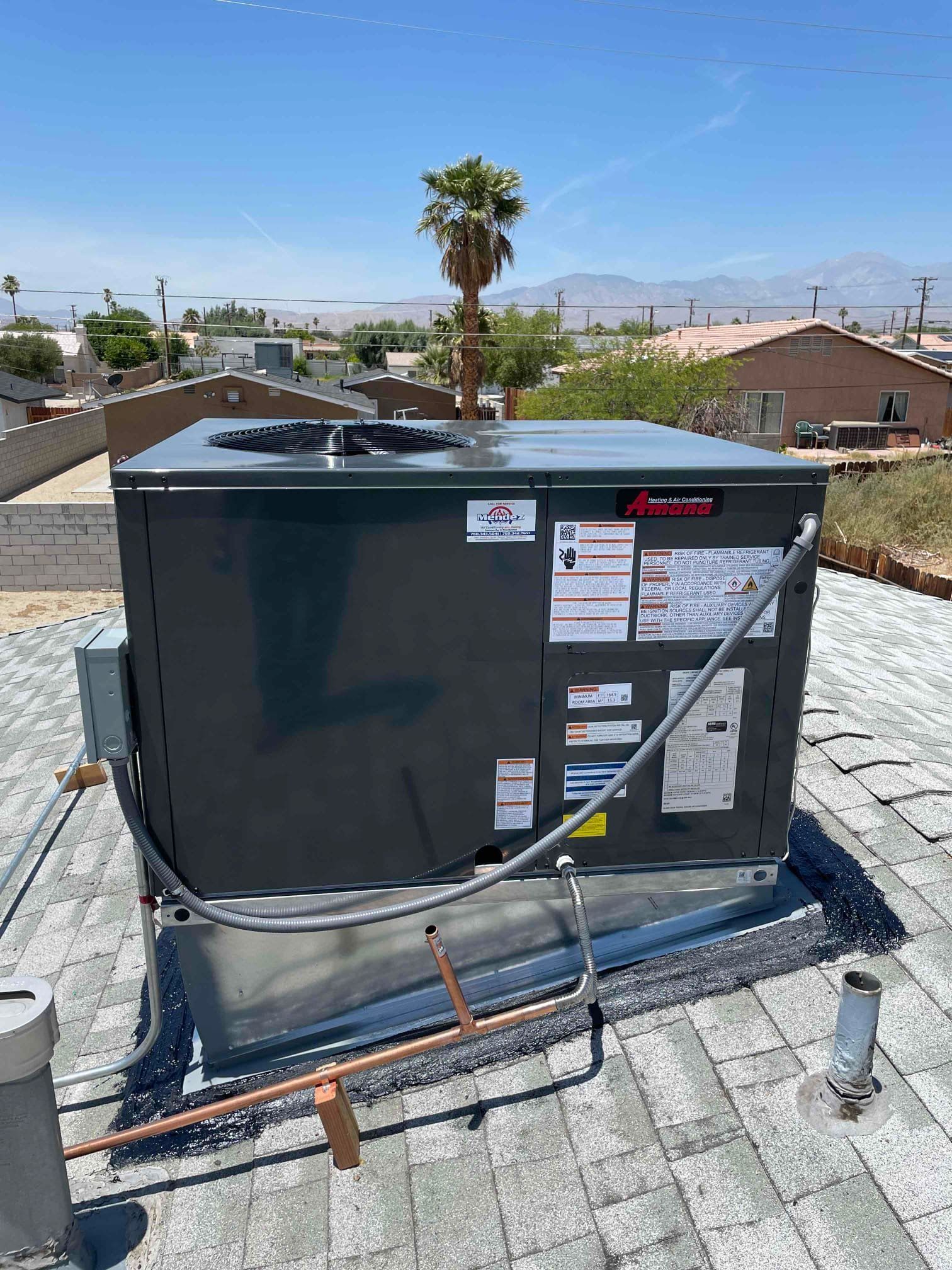 Dark-colored HVAC unit on a rooftop with clear blue sky in background. Palm tree and distant buildings are visible.