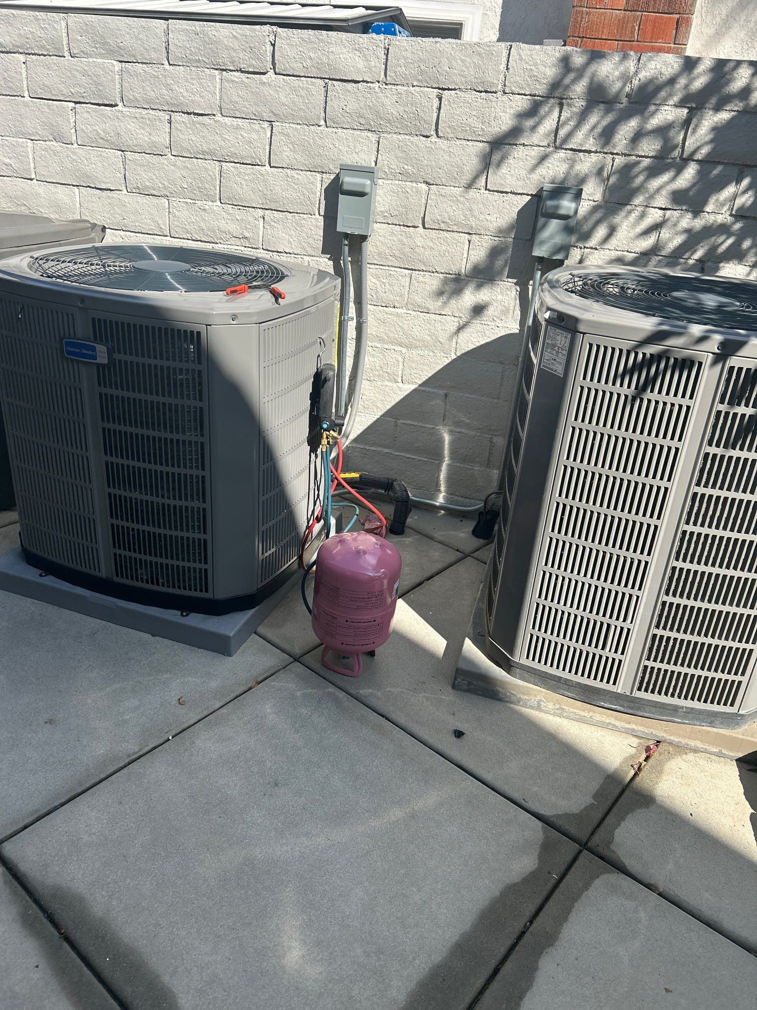 Two air conditioning units outdoors on a concrete pad, connected by tubing to a pink bag.