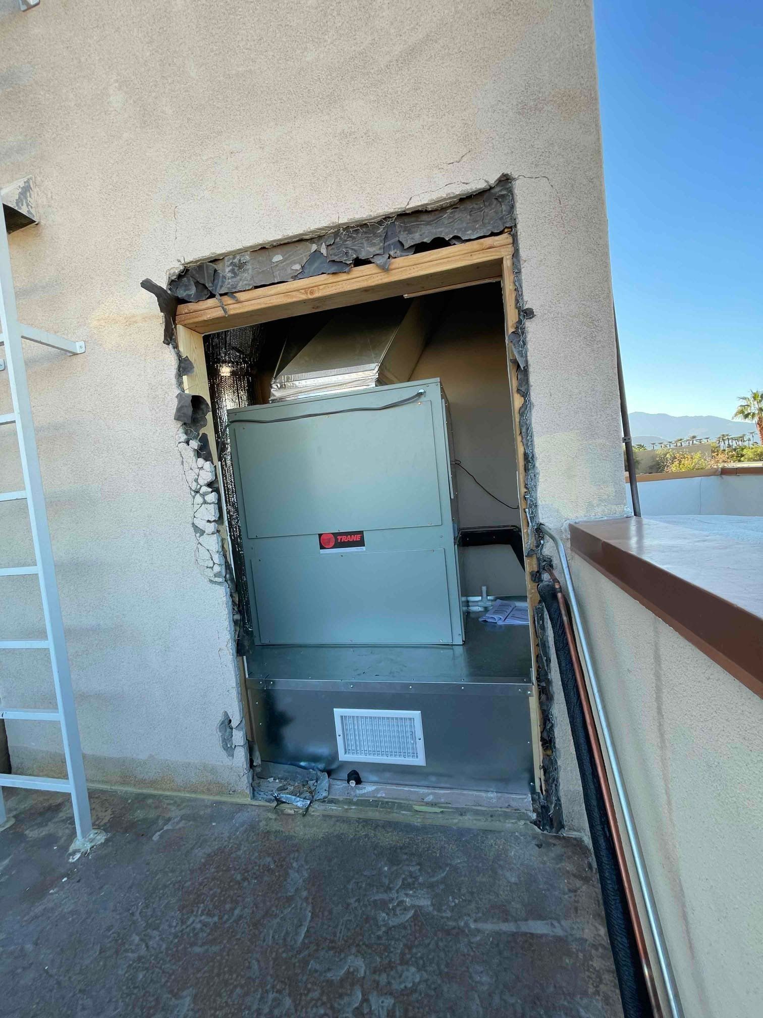 HVAC unit installed in an opening on a rooftop with ductwork visible, against a light-colored stucco wall.