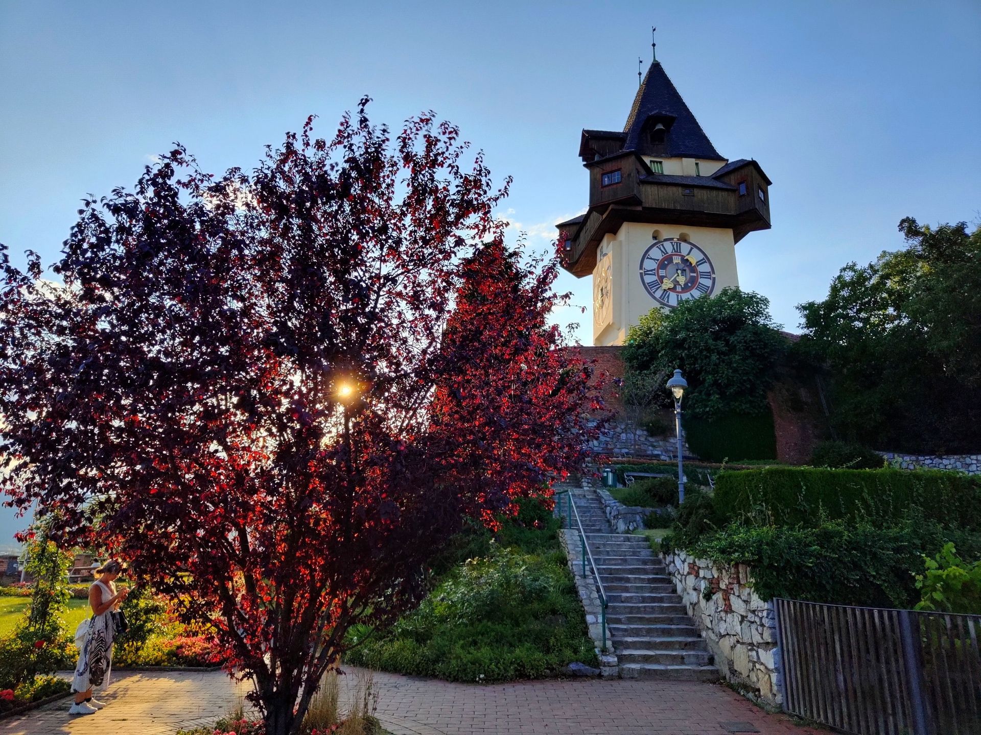 Clock tower overlooking a garden with red-leaved trees, a stone staircase, and a fence; daytime.