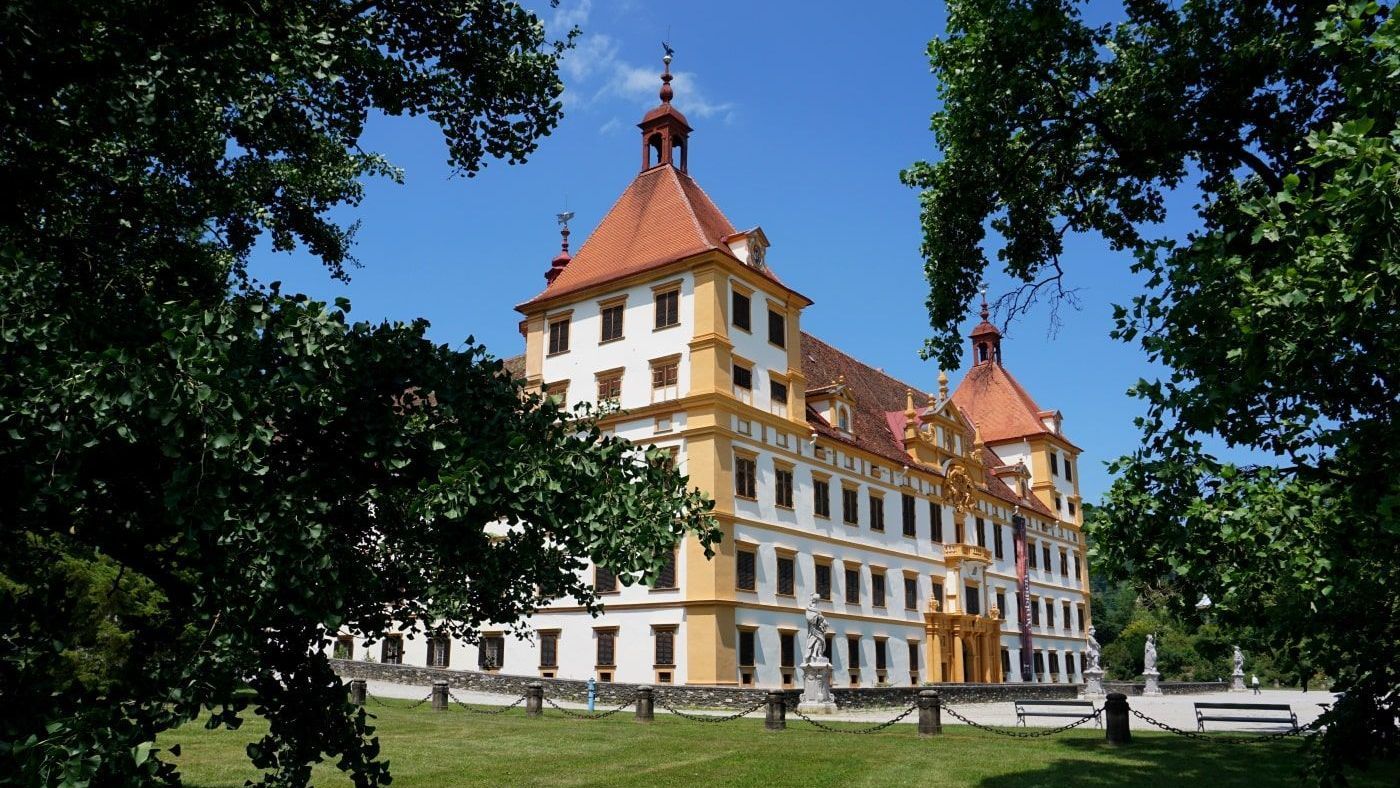 Baroque palace with orange rooftops and a yellow and white facade, framed by green trees against a blue sky.