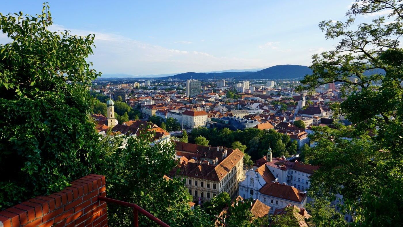 Cityscape of Graz, Austria, viewed from above. Red-tiled roofs, buildings, and greenery are visible under a blue sky.