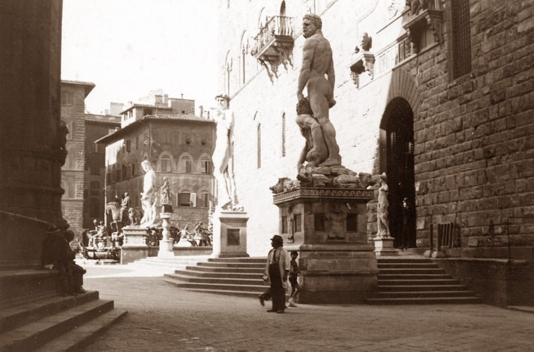 Sepia-toned view of Piazza della Signoria in Florence with statues and pedestrians near Palazzo Vecchio.