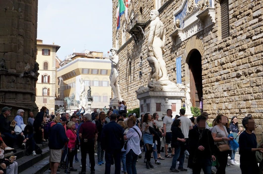 A crowded street scene in Florence, Italy, featuring the Palazzo Vecchio and a large marble statue near the entrance.