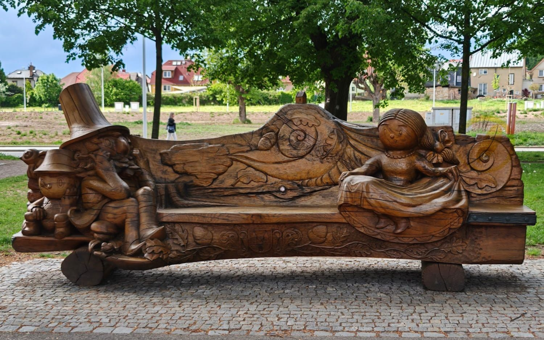 Rust-colored sculpture of a reclining figure on a bench in a park, with trees and a walkway behind.