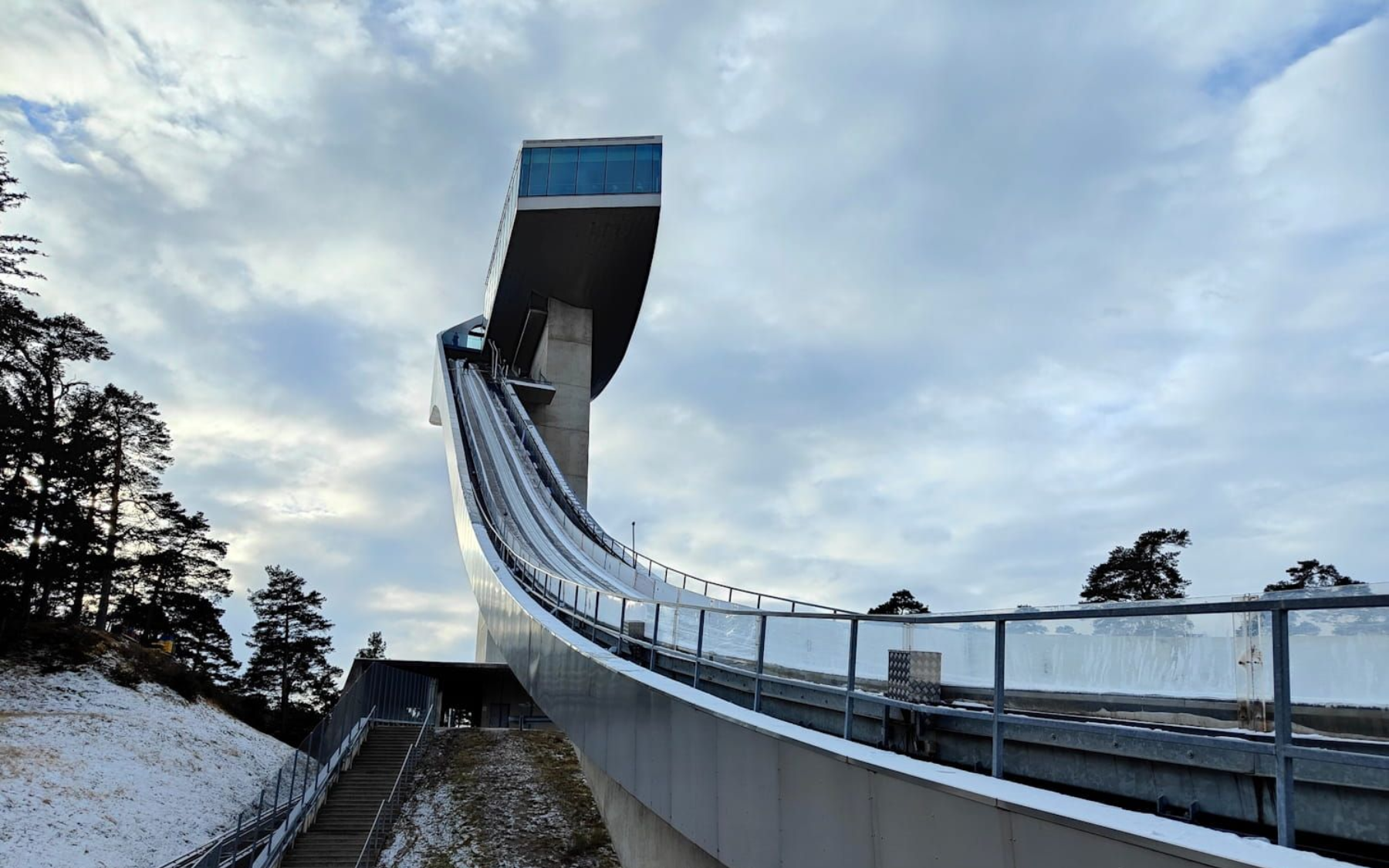 Ski jump ramp in winter, leading to a high tower platform, with cloudy sky.