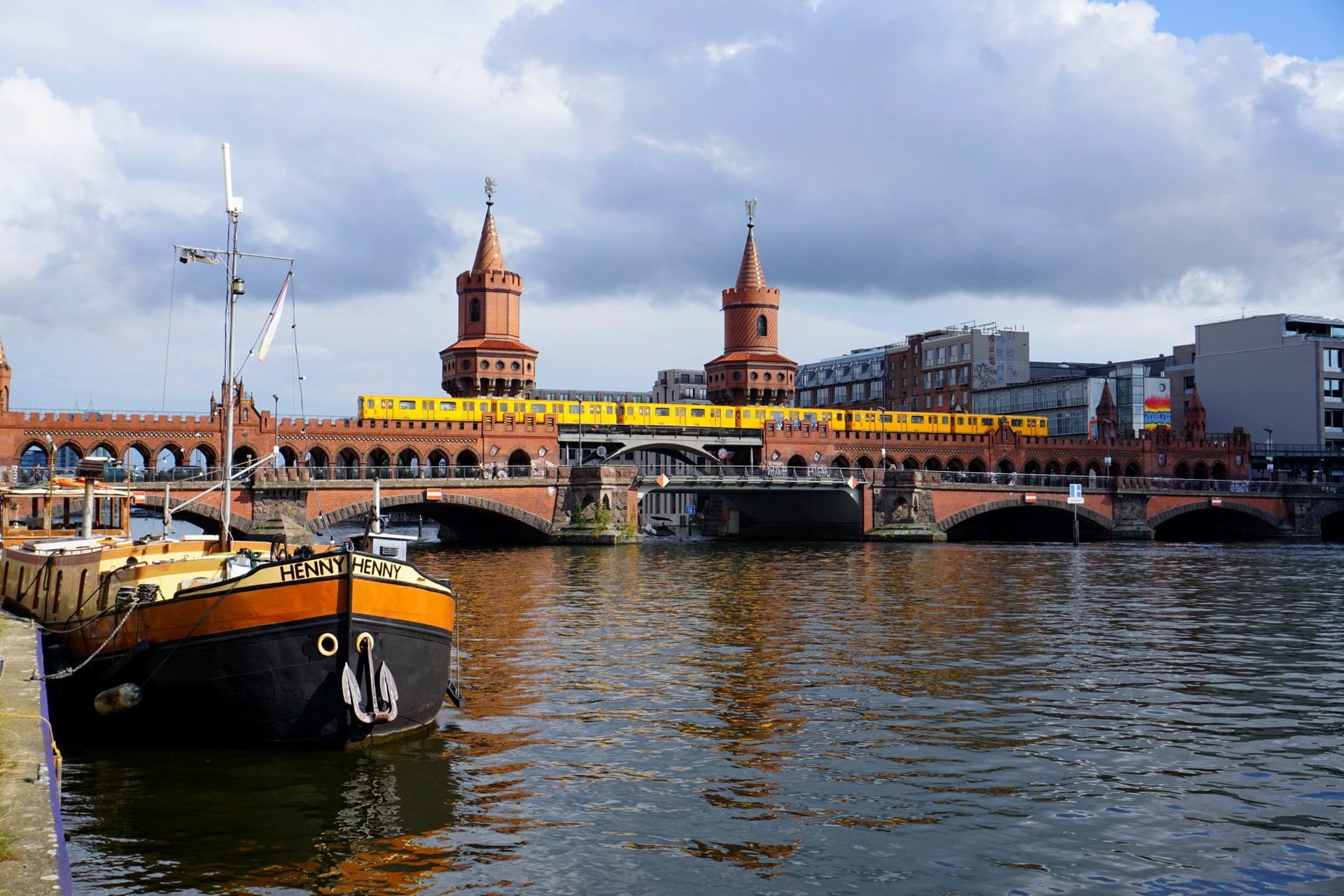Boat in foreground, Oberbaum Bridge in Berlin with train crossing on a cloudy day.