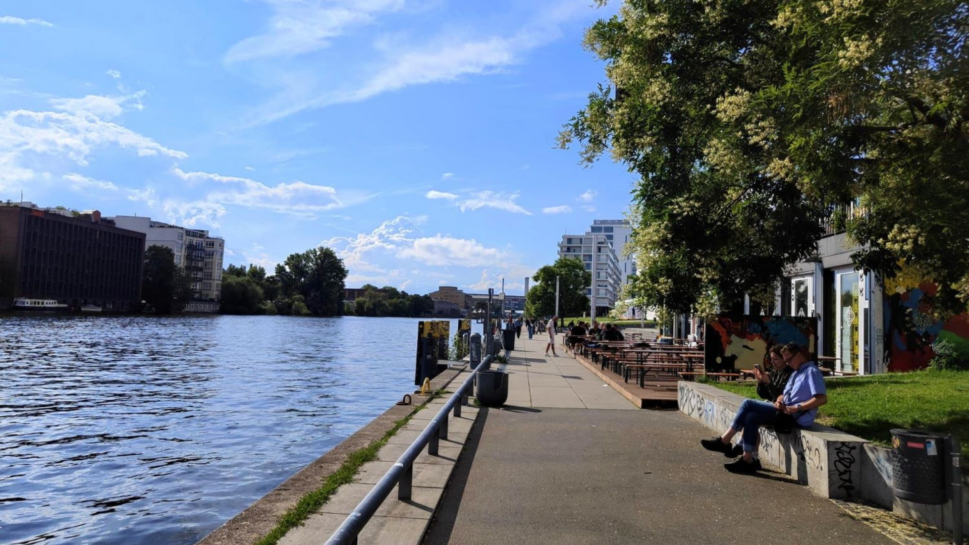 Berlin Spree A sunny day at a waterfront promenade. Man sits, looking at the water, with buildings and blue sky in background.