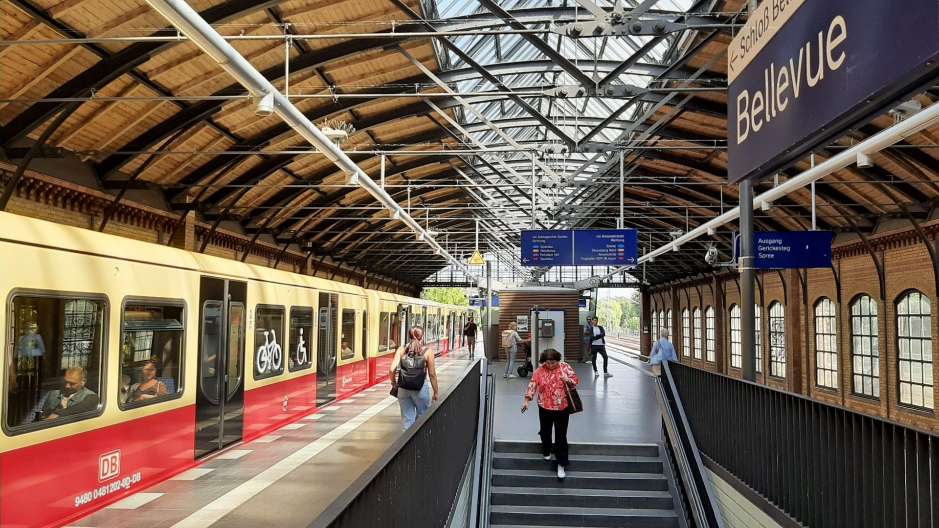 Berlin S-Bhf Bellevue Train at Bellevue station. Passengers walk on platform. Red and yellow train, brick building, sunny day.