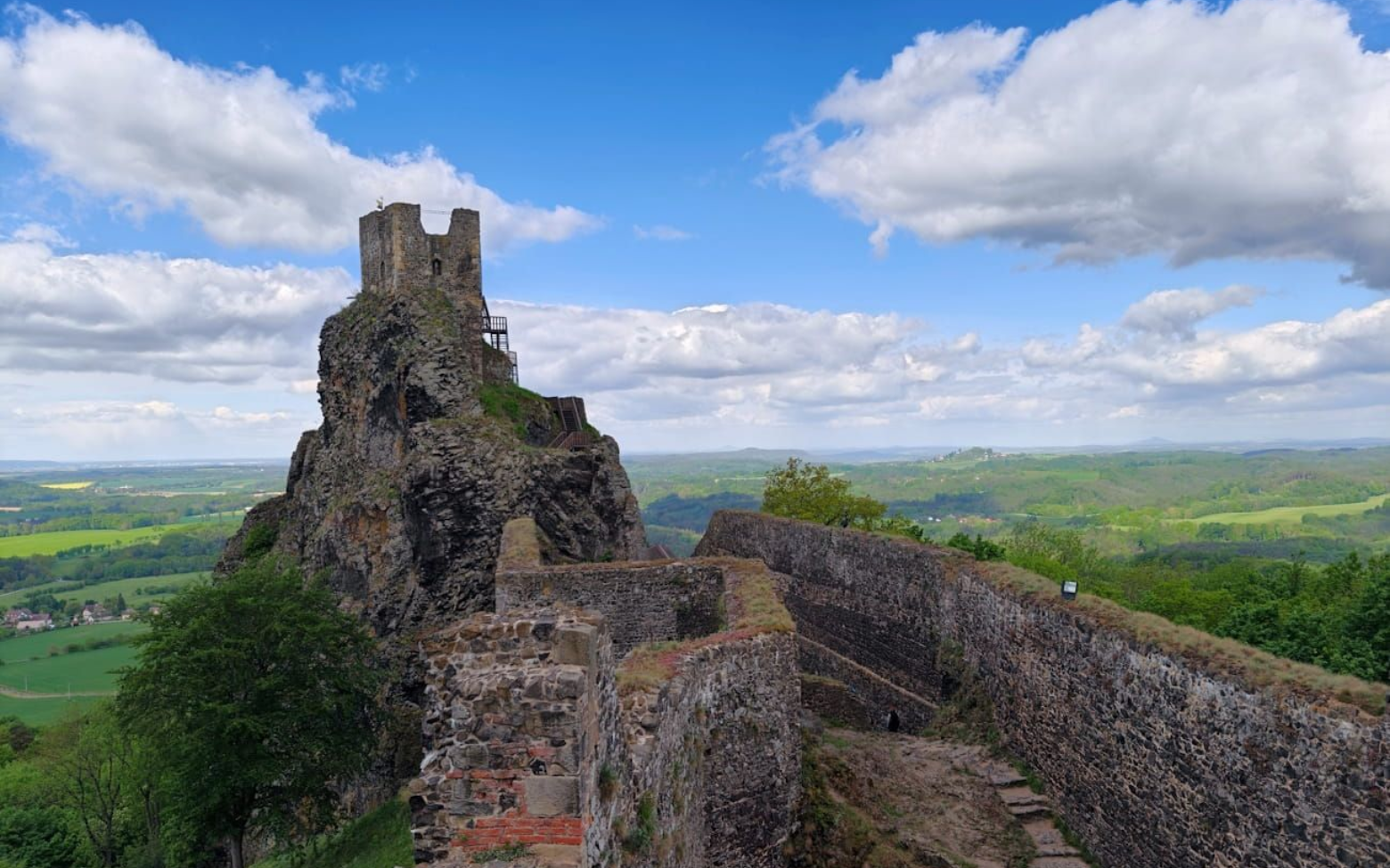 Ruins of a stone castle on a hill, with a winding wall overlooking green countryside under a cloudy sky