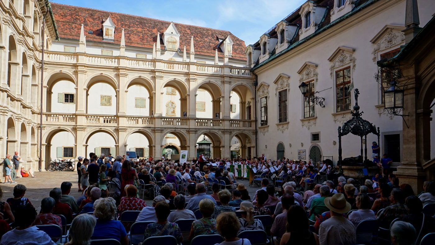People seated in a courtyard, watching a performance. The courtyard has arched walkways and a white building.