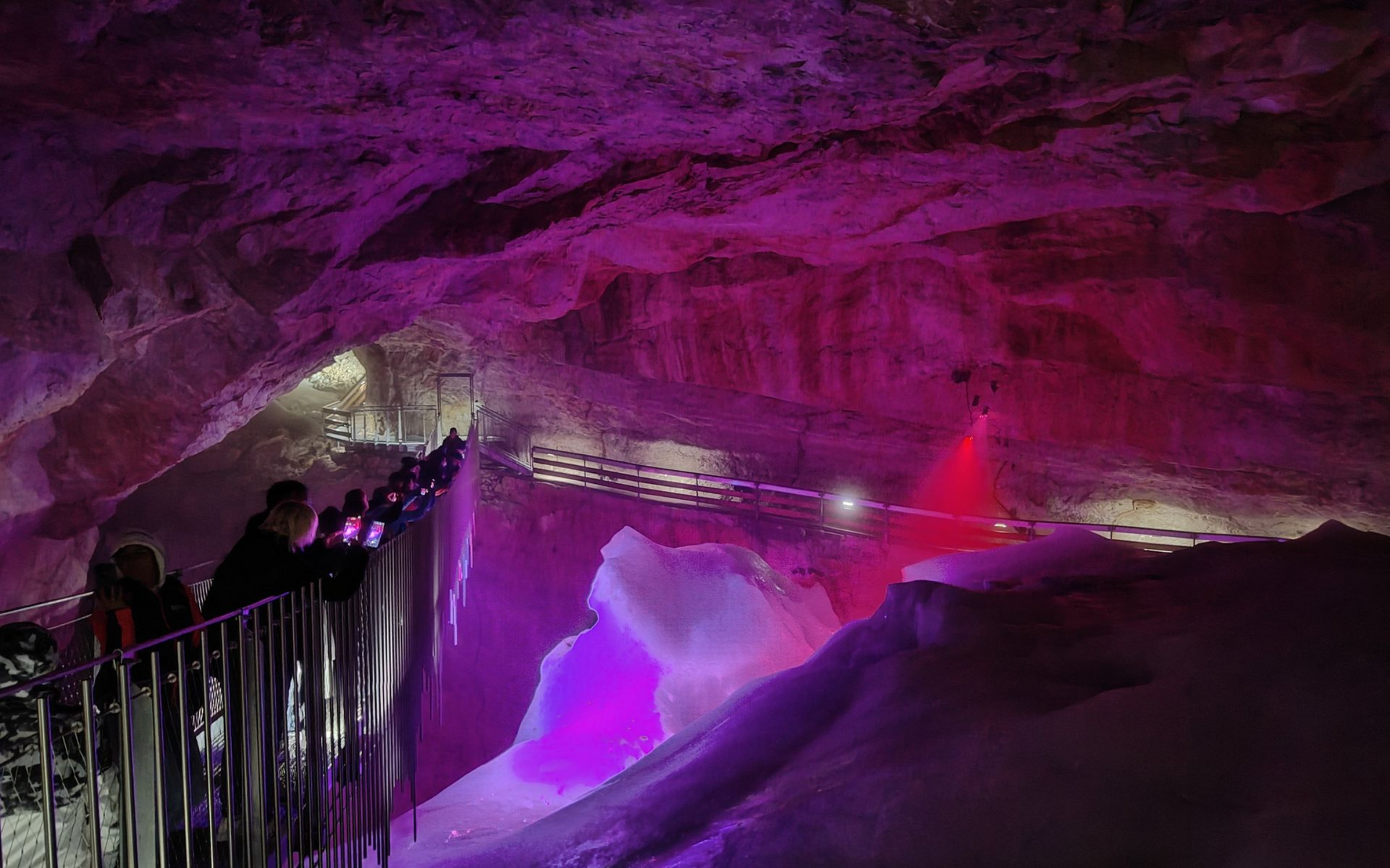Rieseneishöhle Hängebrücke People in a cave with purple and red lighting, ice formations, and a walkway.