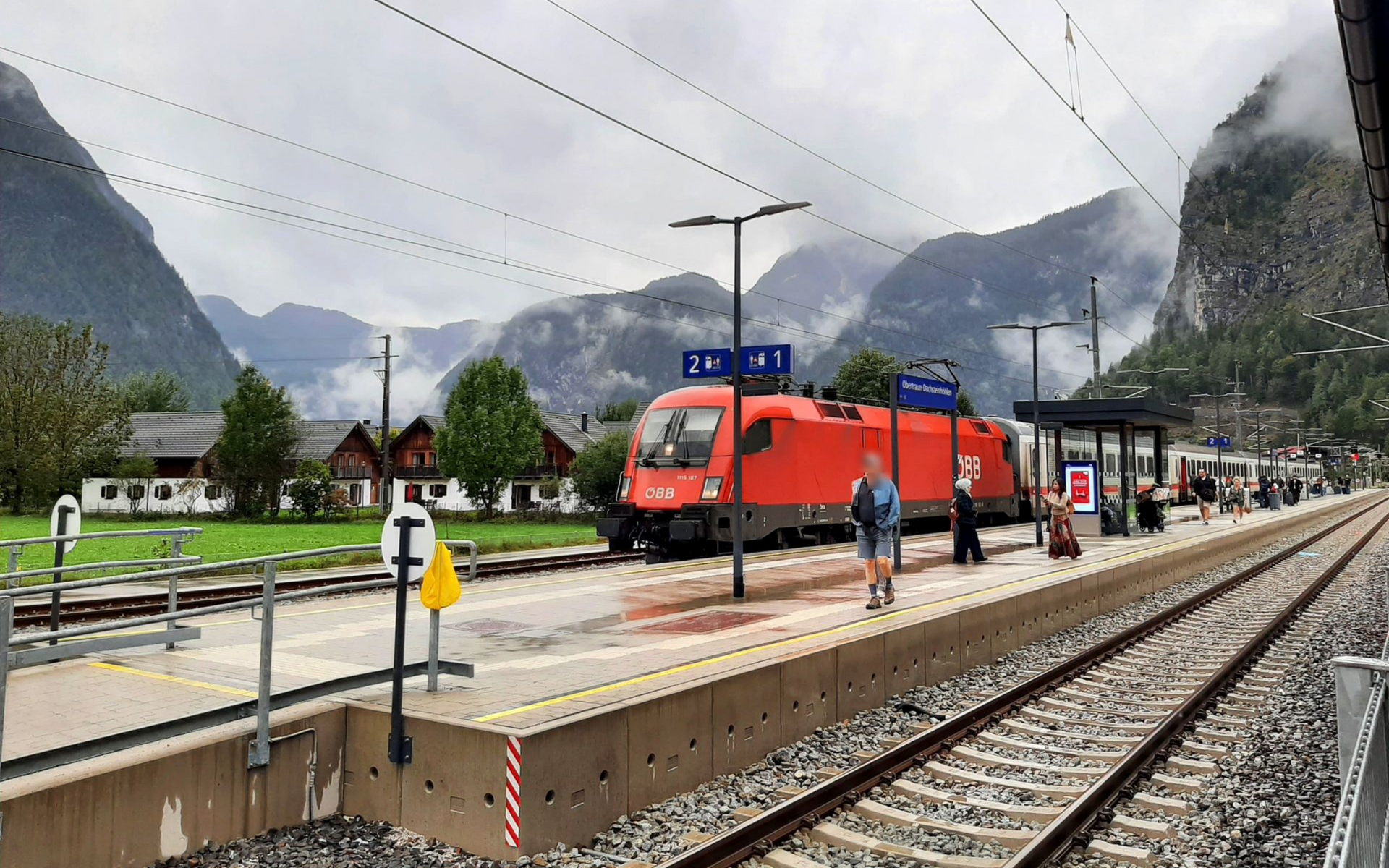 Obertraun-Dachstein A red train at a station platform in a mountainous area, under a cloudy sky.