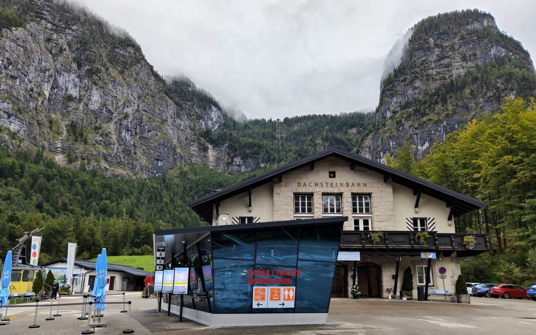Dachstein Krippenstein Eingang A building with a mountain backdrop, blue kiosk, and cars in a parking lot. Cloudy sky.