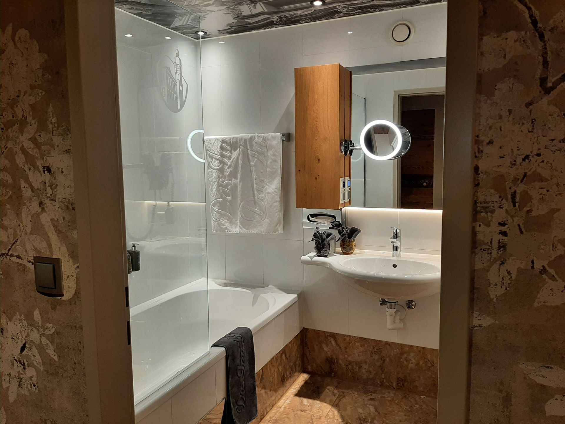 Bathroom with a bathtub, sink, mirror, and wooden cabinet. Beige and white tones, with a decorative floral wall.