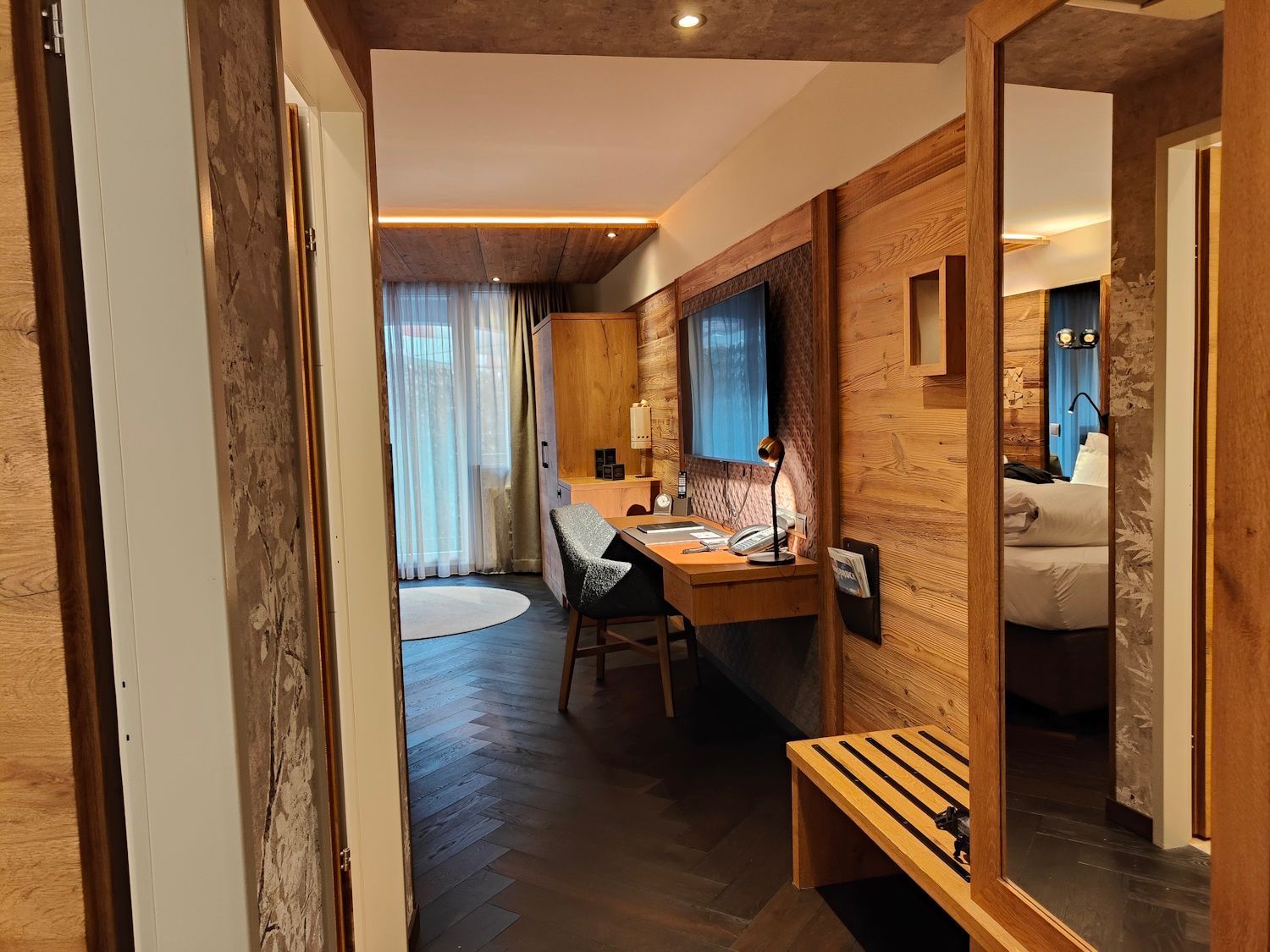 Hotel room interior: wooden walls, desk with chair, TV, hallway with luggage rack, and bed visible in the mirror.