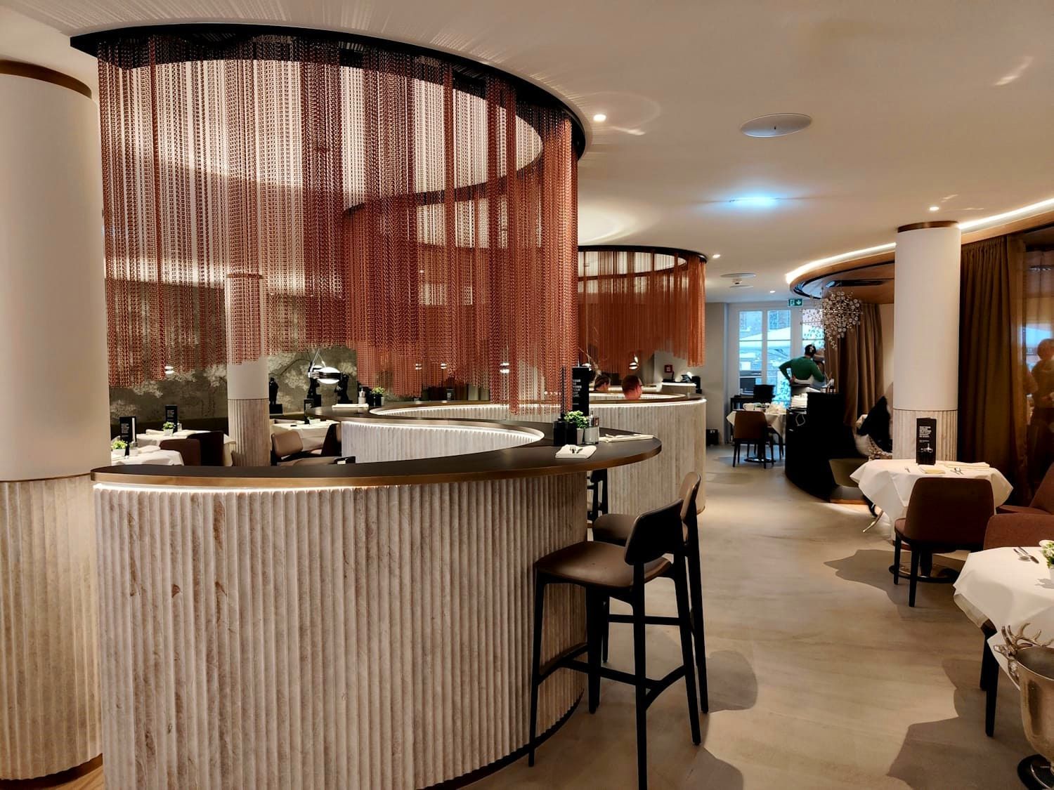 Restaurant interior: bar with wooden facade, hanging orange strands, tables set with white tablecloths.